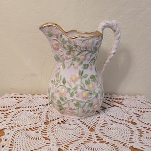 May include: A ceramic pitcher featuring a raised, embossed floral pattern with pink flowers and green vines against a white background. The pitcher has a scalloped rim with a gold-painted edge and a textured, twisted handle extending from the side.