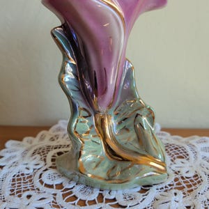 Può includere: Un vaso in ceramica a forma di fiore, con finitura viola e oro. Il vaso ha un'apertura svasata ed è sostenuto da foglie verdi con accenti dorati. La base è di colore verde chiaro.