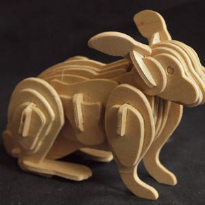 Può includere: Una scultura di coniglio in legno realizzata con pezzi di legno sovrapposti. Il coniglio è in piedi su tutte e quattro le zampe con le orecchie dritte. La scultura è dipinta in un colore legno naturale.
