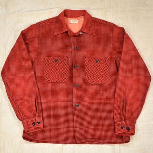 May include: A long-sleeve button-down shirt featuring a red and black micro-check pattern. The shirt includes a pointed collar, two chest pockets with button closures, and buttoned cuffs. The interior collar area shows a light-colored fabric lining.
