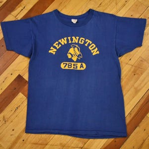 May include: A blue t-shirt with the word "NEWINGTON" arched across the top in yellow letters. Below the text is a yellow Native American head graphic and the numbers "785 A". The shirt is on a wooden surface.