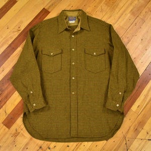 Puede incluir: Camisa abotonada verde oliva con un pequeño estampado de cuadros negros. La camisa tiene dos bolsillos en el pecho, un cuello y puños abotonados. La camisa está tendida sobre una superficie de madera.