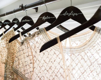 May include: A row of dark wooden hangers with personalized names in white script, holding embellished dresses. The dresses are in shades of cream and gold, with intricate beadwork and sequin details. The scene suggests a bridal or special occasion.