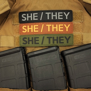May include: Three rectangular patches with the text "SHE / THEY" in black lettering. The patches are black, orange, and olive green. Below the patches are three black magazines.