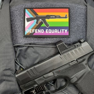 Peut inclure: Un pistolet noir avec le mot "HELLCAT" imprimé sur le côté, et un patch avec un drapeau arc-en-ciel et les mots "DEFEND EQUALITY". Le patch présente une silhouette de fusil noir.