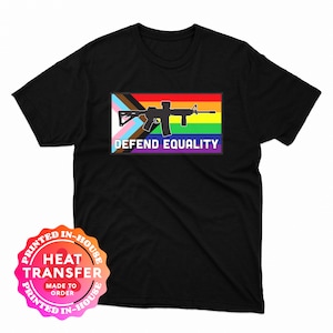 May include: Black t-shirt featuring a graphic with a rifle silhouette over a rainbow flag and the text "DEFEND EQUALITY." The design incorporates the transgender pride flag. The shirt has a crew neck and short sleeves.