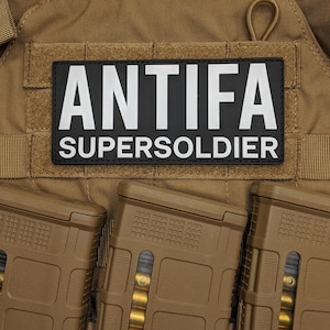 May include: A tan tactical vest with three brown magazines. A black and white patch on the vest reads "ANTIFA SUPERSOLDIER". The vest has a hook and loop patch for attaching accessories.