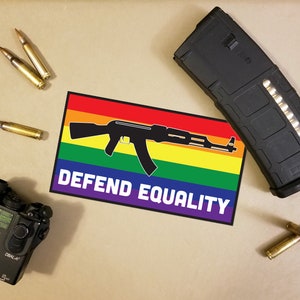 Pode incluir: Um conjunto de equipamentos táticos, incluindo um carregador preto, um dispositivo laser e várias balas. Um adesivo com uma bandeira arco-íris e uma silhueta de rifle preto diz "DEFEND EQUALITY".