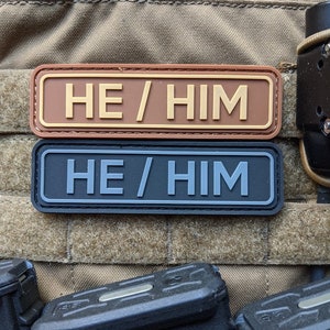 May include: Two rectangular patches with the text "HE / HIM". One patch is brown with tan text, the other is black with grey text. The patches are displayed on a tan tactical vest.