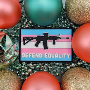 Puede incluir: Un parche rectangular con una silueta de rifle negro sobre un fondo rosa, blanco y azul, con las palabras "DEFEND EQUALITY" en blanco. El parche está rodeado de adornos navideños rojos, dorados y verde azulado, y una guirnalda de cuentas plateadas.