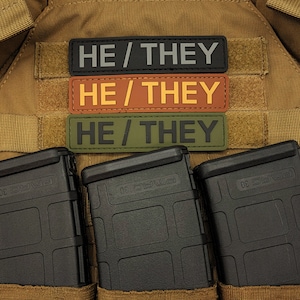 Könnte beinhalten: Drei rechteckige Aufnäher mit dem Text "HE / THEY" in einer serifenlosen Schriftart. Der obere Aufnäher ist schwarz mit grauem Text, der mittlere ist hellbraun mit gelbem Text und der untere ist olivgrün mit schwarzem Text. Die Aufnäher befinden sich auf einem beigen Hintergrund.