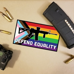 Può includere: Un assortimento di equipaggiamento tattico tra cui una toppa con una bandiera arcobaleno e la sagoma di un fucile, la scritta "DEFEND EQUALITY", un caricatore nero, proiettili e un mirino laser.