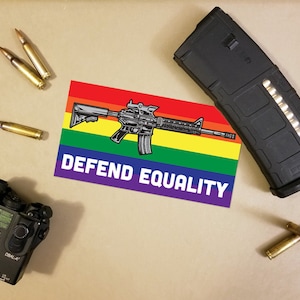 Peut inclure: Divers éléments, dont un chargeur noir, des balles et un autocollant avec un fond arc-en-ciel et un fusil. L'autocollant indique "DEFEND EQUALITY". Un appareil laser noir est également présent.