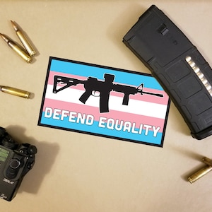 May include: An assortment of tactical gear including a black rifle magazine, a laser device, and several bullets. A sticker with a rifle silhouette over the transgender flag and the words "DEFEND EQUALITY" is also present.