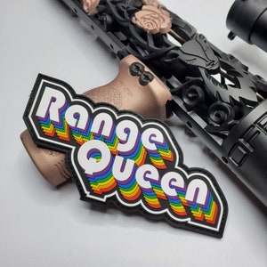 May include: A black and white patch with the text "Range Queen" in a rainbow design. The patch is placed on a rose gold coloured rifle grip, with a black rifle barrel in the background.