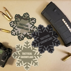 May include: An assortment of tactical gear, including a black magazine, a laser device, and several bullets. Four snowflake-shaped stickers with the text "TACTICAL SNOWFLAKE" in various shades of gray and camouflage are also present.