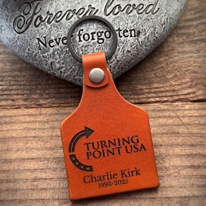 May include: Orange leather keychain with a black metal ring. The keychain features the text "TURNING POINT USA" and "Charlie Kirk 1993-2025". The keychain is on a wooden surface.