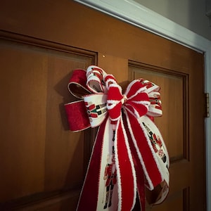 May include: A large, decorative Christmas bow with red and white striped velvet ribbon and a patterned ribbon featuring toy soldiers. The bow is attached to a brown wooden door, creating a festive holiday decoration.