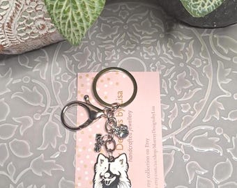 May include: A silver keychain with a dog charm, a paw print charm, and a small charm that says "love". The dog charm is a black and white illustration of a dog. The keychain is attached to a pink card with text.