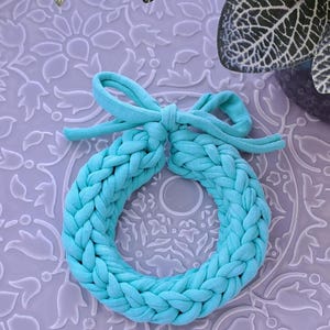 Könnte beinhalten: Ein geflochtener Stoffkranz in Türkisblau mit Schleife. Der Kranz ist aus weichem, gewebtem Stoff gefertigt und perfekt, um jedem Raum einen Hauch von Farbe zu verleihen.