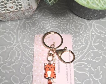 May include: A gold-colored keychain with a cartoon cat charm. The orange and brown cat has a smiling face and striped pattern. The keychain includes a split ring and a lobster clasp. A pink card with text is attached.