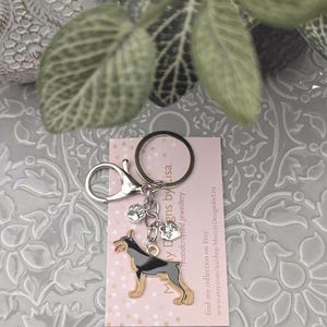May include: A silver keyring featuring a Doberman Pinscher charm. The charm is black and tan with a red tongue. Includes a paw print and heart charm. The pink tag reads "May Day Designs by Lisa" and "handcrafted jewellery".