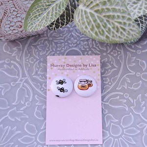 Puede incluir: Pendientes de botón blancos con diseños de abeja y tarro de miel sobre una tarjeta rosa. La tarjeta tiene el texto "Murray Designs by Lisa" y "Handcrafted in Adelaide". El fondo incluye hojas verdes y una superficie decorativa.