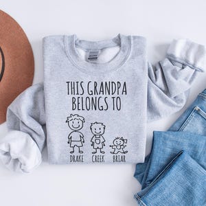 Op de afbeelding: Grijs sweatshirt met de tekst "This Grandpa Belongs To" en drie stick figuren met de namen "Drake", "Creek" en "Briar" eronder.