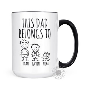 May include: White ceramic coffee mug with a black rim and handle. The mug has black text that reads "This Dad Belongs To" with stick figure drawings of three children and their names: Logan, Gavin, and Nora.