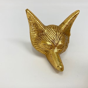 Könnte beinhalten: Eine goldfarbene Metallskulptur eines Fuchs Kopfes. Der Fuchs hat ein detailliertes Gesicht mit einer langen Schnauze und spitzen Ohren.