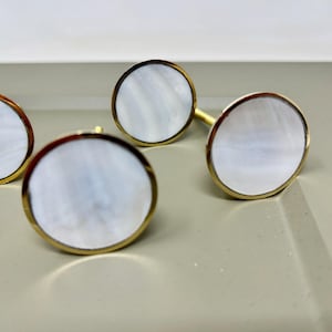 May include: Four circular cabinet knobs featuring a mother-of-pearl centre and a gold-coloured rim. The knobs are displayed on a light grey surface, highlighting their elegant design. These decorative knobs are suitable for furniture enhancements.