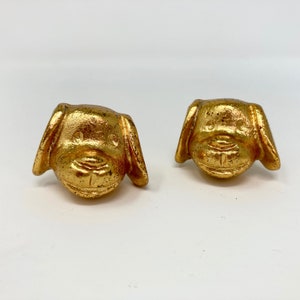 Puede incluir: Un par de pendientes de cabeza de perro de color dorado. Los pendientes son pequeños y tienen un diseño detallado.