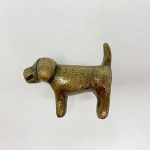 Könnte beinhalten: Eine goldfarbene Metallfigur eines Hundes. Der Hund steht auf allen vieren mit eingerolltem Schwanz. Die Figur ist schlicht gehalten und hat eine glatte Oberfläche.