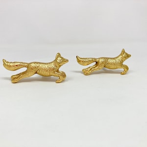 May include: Two gold-coloured fox figurines, each running in a different direction. The foxes are detailed with textured fur and have a realistic appearance.