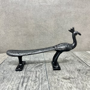 May include: Black metal peacock door handle with intricate details. The handle is shaped like a peacock with its tail feathers spread out. It has two mounting plates for installation.