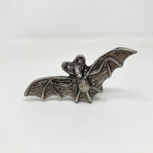 Könnte beinhalten: Eine silberne Fledermaus-Figur aus Metall mit detaillierten Flügeln und einer strukturierten Oberfläche.