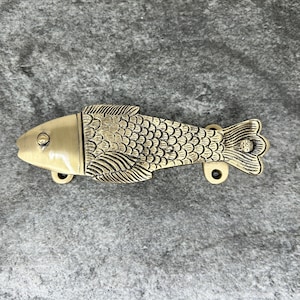 May include: A gold-toned fish-shaped door handle with a detailed design of scales and fins. The handle is mounted on a gray background.