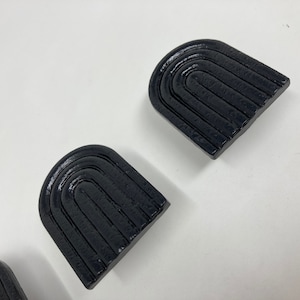 Może przedstawiać: Dwa czarne, łukowate uchwyty do szuflad z fakturowaną powierzchnią. Uchwyty mają zaokrąglony górny brzeg i płaskie dno.