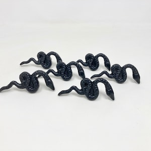 Puede incluir: Cinco figuritas de serpiente de plástico negro, cada una con un cuerpo enroscado y una cola puntiaguda. Las serpientes están dispuestas en semicírculo, con las cabezas mirando hacia adentro.