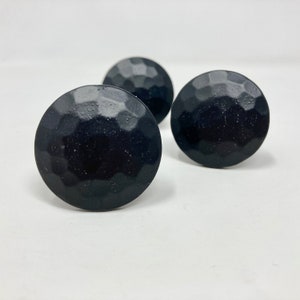 Könnte beinhalten: Drei schwarze, gehämmerte Metallknöpfe mit runder, leicht gewölbter Form.