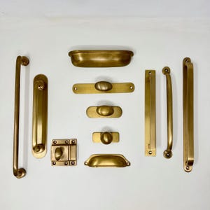 May include: A selection of antique brass hardware, including handles, pulls, and a latch. The hardware pieces vary in size and shape, with a warm, golden hue. The arrangement showcases a variety of designs suitable for furniture or cabinetry.