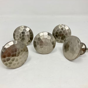 Peut inclure: Cinq boutons de cabinet en métal martelé argenté. Les boutons sont ronds et ont une surface texturée.