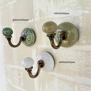 Può includere: Tre ganci da parete in ceramica con ganci in ottone anticato e pomelli in ceramica. I pomelli sono verdi, verde oliva e bianchi. Il testo "green/antique", "olive/antique" e "white/antique" è scritto sotto ogni gancio.