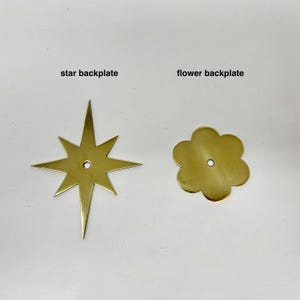 Peut inclure: Deux plaques arrière en laiton : une en forme d'étoile et une en forme de fleur. La plaque arrière en étoile a huit pointes. La plaque arrière en fleur a un bord arrondi et festonné. Les deux ont un trou central et le texte "star backplate" et "flower backplate".
