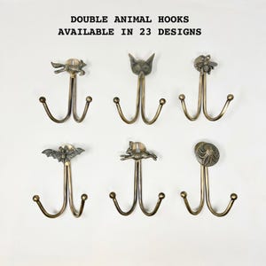 May include: Six double animal hooks in a bronze finish are displayed against a white background. The hooks feature various animal designs, including a bat, bee, fox, and rabbit. The text "DOUBLE ANIMAL HOOKS AVAILABLE IN 23 DESIGNS" is at the top.