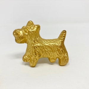 Puede incluir: Una figurita de metal de color dorado de un perro terrier. La figurita está detallada con un patrón de pelaje texturizado.