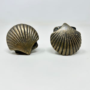 Może przedstawiać: Dwa uchwyty do szafek w kształcie muszli w kolorze brązu. Każdy uchwyt ma szczegółowy, żebrowany wzór, przypominający przegrzebek. Uchwyty są umieszczone na gładkim, białym tle, podkreślając ich skomplikowaną fakturę i kształt.
