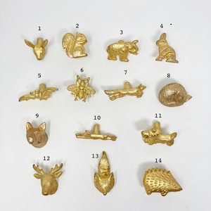 May include: Set of 14 gold-toned animal and nature-themed cabinet knobs. The knobs feature a variety of designs, including a squirrel, a fox, a deer, a bee, a hedgehog, a rabbit, a bat, a branch, and a snail.