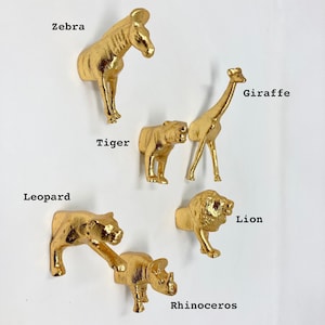 May include: A collection of six gold-colored animal figurines: a zebra, tiger, giraffe, leopard, rhinoceros, and lion. Each figurine is mounted on a white surface, with the animal's name printed in black text nearby.
