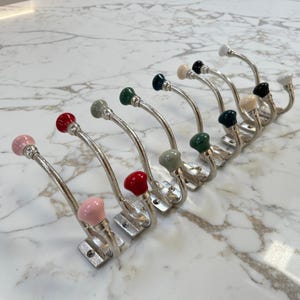 May include: A set of decorative metal coat hooks with ceramic knobs in various colours. The knobs are pink, red, green, grey, black, and cream. The hooks have a curved design and are mounted on a rectangular base.